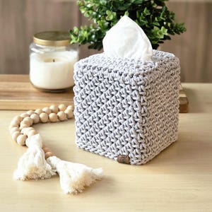 May include: A gray, crocheted tissue box cover with a square shape. The cover has a textured, woven appearance. White tissues are visible at the top. A wooden bead garland with tassels and a candle in a jar are in the background.