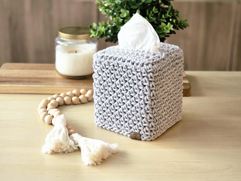 May include: A gray, crocheted tissue box cover with a square shape. The cover has a textured, woven appearance. White tissues are visible at the top. A wooden bead garland with tassels and a candle in a jar are in the background.