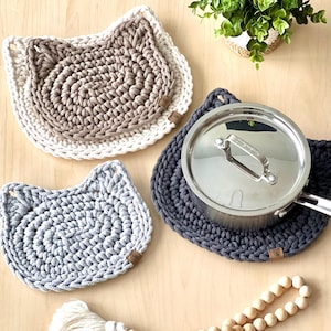 May include: Three crocheted cat-shaped pot holders in grey, beige, and white. The pot holders are on a wooden surface with a wooden bead garland and a potted plant.
