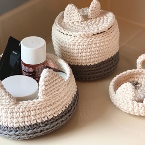 May include: Three crocheted cat-shaped baskets with gray and white yarn. The largest basket has a lid and is filled with toiletries. The smallest basket has a handle and is filled with jewelry.