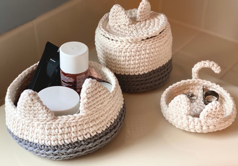May include: Three crocheted cat-shaped baskets with gray and white yarn. The largest basket has a lid and is filled with toiletries. The smallest basket has a handle and is filled with jewelry.