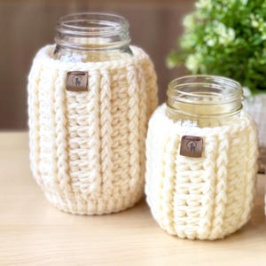 May include: Two clear glass jars with cream-coloured, chunky knit cosies. Each cosy has a small, brown leather tag. The jars are on a light-coloured wooden surface. The cosy is a decorative item.