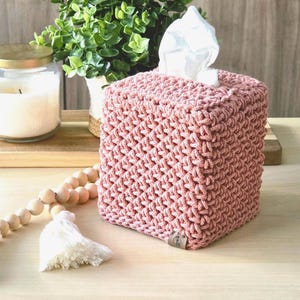 May include: A square, dusty rose macrame tissue box cover with a textured pattern. White tissues peek out from the top. A candle in a glass jar and a wooden bead garland with tassels are in the background. Home decor.