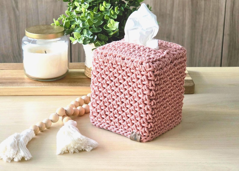 May include: A square, dusty rose macrame tissue box cover with a textured pattern. White tissues peek out from the top. A candle in a glass jar and a wooden bead garland with tassels are in the background. Home decor.
