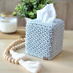 May include: A light blue, square tissue box cover with a textured, woven design. White tissues peek out from the top. A wooden bead garland with white tassels and a lit candle in a glass jar are in the background.