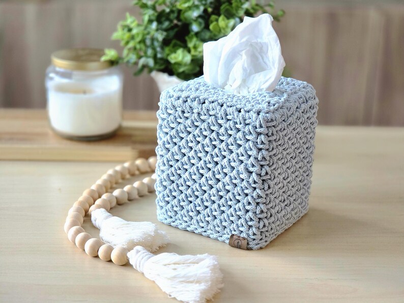 May include: A light blue, square tissue box cover with a textured, woven design. White tissues peek out from the top. A wooden bead garland with white tassels and a lit candle in a glass jar are in the background.