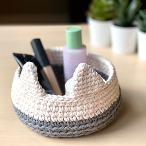 May include: A white and gray crocheted bowl with a crown-like rim. The bowl is filled with various toiletries, including a bottle of lotion, a tube of cream, and a makeup brush.