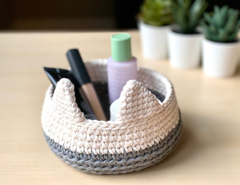 May include: A white and gray crocheted bowl with a crown-like rim. The bowl is filled with various toiletries, including a bottle of lotion, a tube of cream, and a makeup brush.