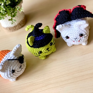 Puede incluir: Tres juguetes de gato de crochet disfrazados para Halloween. Uno es una bruja con un sombrero negro y un cuerpo verde, otro es un vampiro con una capa roja y un cuerpo blanco, y el &uacute;ltimo es un pirata con un cuerpo naranja y blanco y un parche en el ojo.