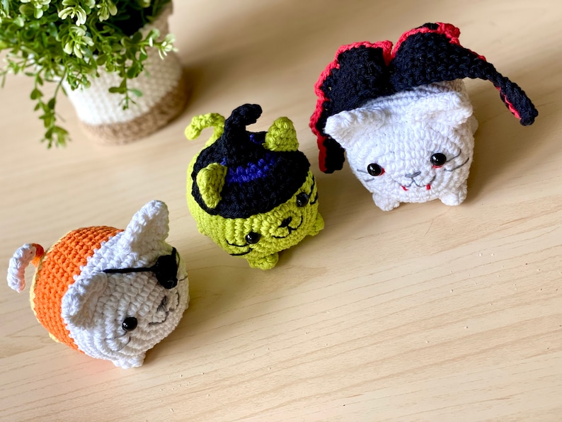 Puede incluir: Tres juguetes de gato de crochet disfrazados para Halloween. Uno es una bruja con un sombrero negro y un cuerpo verde, otro es un vampiro con una capa roja y un cuerpo blanco, y el &uacute;ltimo es un pirata con un cuerpo naranja y blanco y un parche en el ojo.