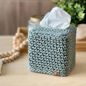 May include: A square, sage green tissue box cover with a textured, woven design. The cover is made of a soft material and has a small, brown leather tag on the bottom corner. White tissues are visible at the top.