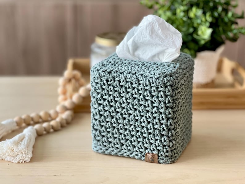 May include: A square, sage green tissue box cover with a textured, woven design. The cover is made of a soft material and has a small, brown leather tag on the bottom corner. White tissues are visible at the top.