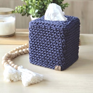 May include: A square, navy blue tissue box cover with a textured, woven design. White tissues peek out from the top opening. A wooden bead garland with white tassels and a candle in a glass jar are also visible.