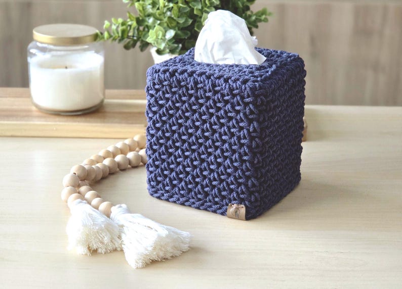 May include: A square, navy blue tissue box cover with a textured, woven design. White tissues peek out from the top opening. A wooden bead garland with white tassels and a candle in a glass jar are also visible.