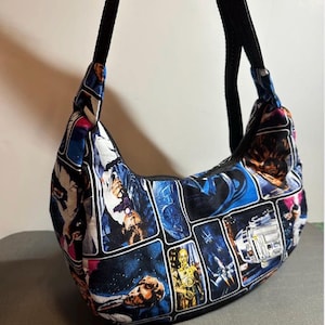Op de afbeelding: Een hobo-stijl schoudertas met een stoffen print van meerdere panelen met Star Wars-personages en scènes, waaronder C-3PO en R2-D2. De tas heeft een zwarte ritssluiting aan de bovenkant en een verstelbare zwarte stoffen schouderband.