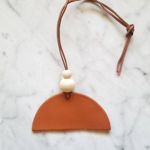 May include: A terracotta half-circle ceramic ornament with a brown leather cord and two white wooden beads.