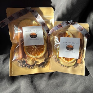 May include: Two tan pouches containing dried orange slices, apple slices, cinnamon sticks, and cloves. Each pouch features a white label with the text Witches Brew Simmer Pot and a graphic of a cauldron, tied with a grey ribbon with owl patterns.