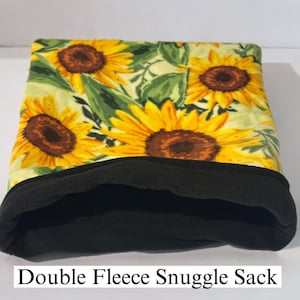 Puede incluir: Un saco de dormir de doble forro polar con un patrón de girasoles de pétalos amarillos y centros marrones sobre un fondo verde claro, contrastado por un forro interior negro sólido. El texto Double Fleece Snuggle Sack es visible.