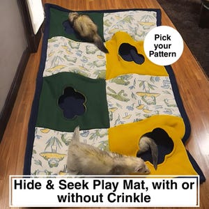 May include: A patchwork play mat with a navy blue border, featuring green and yellow sections with cut-out holes. The mat has a pattern of frogs and mushrooms. Two ferrets are playing on the mat. Text on the mat says "Hide & Seek Play Mat, with or without Crinkle."
