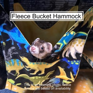 May include: A fleece bucket hammock featuring a black background with a colorful dinosaur print in shades of blue, yellow, and white. The text "Fleece Bucket Hammock" is visible at the top, and a small ferret is peeking out from the center.