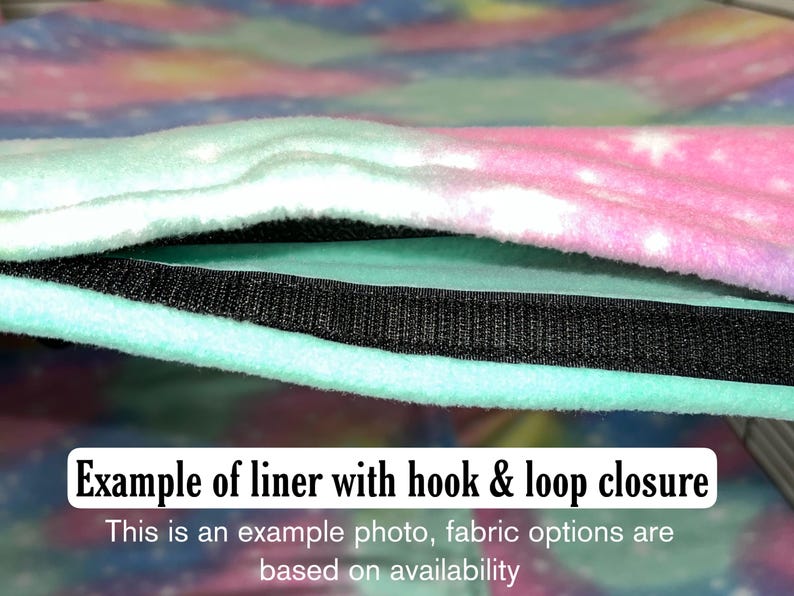 Puede incluir: Un forro de tela suave con un estampado de galaxia en tonos pastel rosa, azul y morado con estrellas blancas. El forro incluye una tira de cierre de velcro negra a lo largo del borde. El texto dice: Example of liner with hook & loop closure.
