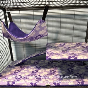 Puede incluir: Una hamaca de forro polar para mascotas con estampado floral morado y blanco, una funda de plataforma rectangular a juego y una funda de rampa dentro de una jaula de alambre. El texto en la imagen dice: This is an example photo. Please see Item Details.