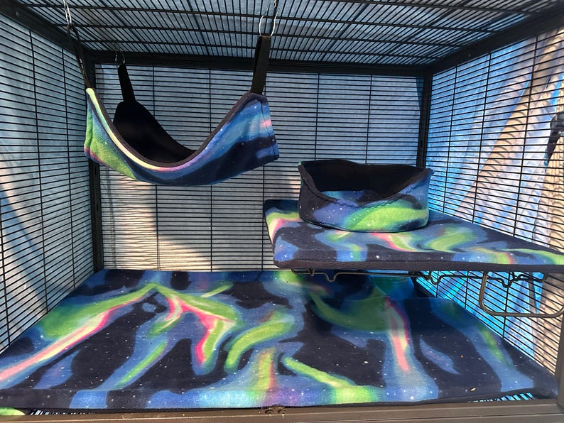 Puede incluir: Un conjunto de accesorios para jaula de animales peque&ntilde;os que incluye una hamaca colgante, una alfombrilla rectangular y una plataforma elevada con cama circular, hechos de tela suave con patr&oacute;n de aurora boreal y bordes negros.