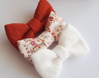 May include: Three fabric hair bows in a set. One bow is solid rust orange, one is white, and one is a floral print with rust, white, and pink flowers.