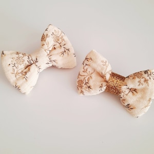 May include: Two beige fabric hair bows with a floral pattern. One bow has a gold glitter accent.