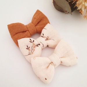 May include: Three fabric hair bows. One bow is a solid brown color. The other two bows are white with a floral pattern and gold glitter accents.