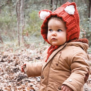 Peut inclure: Un tout-petit portant une veste marron et un bonnet de renard en crochet rouge avec des oreilles blanches. L'enfant tient une petite branche dans sa main et se tient dans une zone boisée.