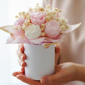 Könnte beinhalten: Ein Blumenstrauß aus rosa und weißen Rosen und kleinen gelben Blüten, präsentiert in einem weißen zylindrischen Behälter. Die Blumen sind auf rosa Seidenpapier angeordnet.