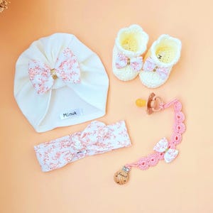 以下が含まれることがあります： ピンクの花柄のリボンが付いた白いターバン、ピンクの花柄のヘッドバンド、ピンクのリボンが付いた白いクロシェのブーティ、そして「Minuk」と書かれた木製のチャームが付いたピンクのクロシェの哺乳瓶クリップ。