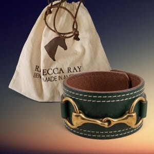 May include: A dark green leather cuff bracelet featuring a brass horse bit hardware detail and white contrast stitching. The bracelet is accompanied by a cream-colored drawstring pouch with a brown horse head logo and the text REBECCA RAY.