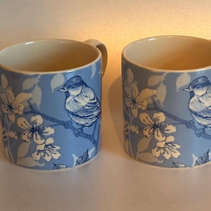 Peut inclure: Un ensemble de quatre tasses en céramique présentant un fond bleu clair avec un motif répétitif de petits oiseaux perchés sur des branches fleuries. Les oiseaux et les fleurs sont dessinés dans un style bleu foncé et blanc, intérieur crème.