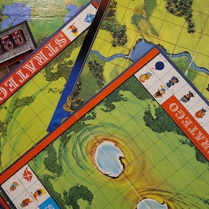 May include: Overhead shot of a Stratego board game. The game board features a green and yellow landscape with grid lines, water features, and the word "STRATEGO" in red. Game pieces are visible.