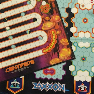 Peut inclure: Un jeu de société vintage avec deux plateaux de jeu, l'un pour Centipede et l'autre pour Zaxxon. Le plateau Centipede est violet avec des chemins orange et blancs et des champignons. Le plateau Zaxxon est noir avec des tuiles hexagonales bleues et blanches.