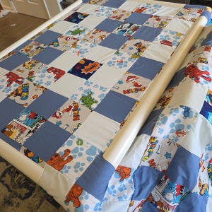 May include: A patchwork quilt with a variety of colorful squares, including blue, white, and patterned fabrics featuring cartoon characters. The quilt is on a white frame, and the design includes paw prints and comic book style graphics.