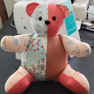 May include: A handmade patchwork teddy bear with a split design. One side features a floral pattern, the other a solid coral color. The bear has black button eyes and is made of various fabrics.
