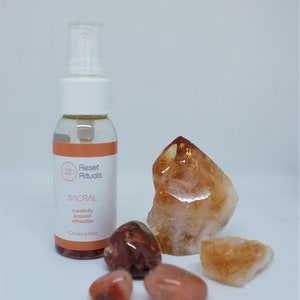 May include: A clear glass spray bottle with a white cap and an orange label that reads "Reset Rituals SACRAL Chakra Mist". The image also features several orange and brown crystals and stones.