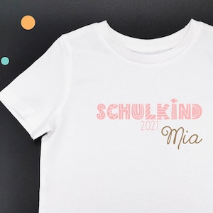 May include: White t-shirt with pink text that says "SCHULKIND 2021 Mia" in a retro font.