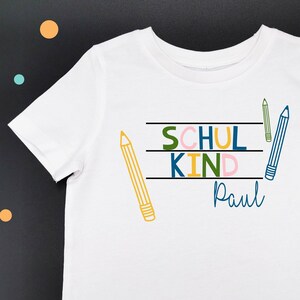 Könnte beinhalten: Weißes T-Shirt mit einer bunten Grafik, auf der "Schul Kind" mit dem Namen "Paul" darunter steht. Die Grafik enthält drei Bleistifte.