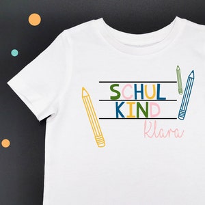 Puede incluir: Camiseta blanca con un gráfico colorido que dice "Schul Kind" con el nombre "Klara" debajo. El gráfico está rodeado de lápices de colores.