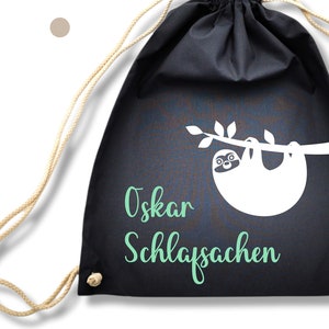 May include: Black drawstring bag with a white sloth hanging from a branch. The text "Oskar Schlafsachen" is written in green on the bag.