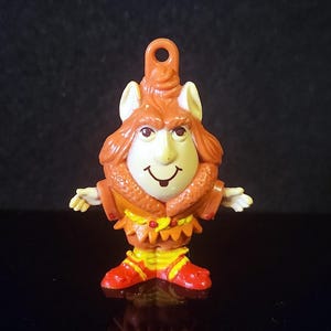 May include: A small, vintage figurine of a cartoon character with a smiling face, wearing a brown hat and outfit with red and yellow accents. The figurine has a loop at the top for hanging. The background is black.