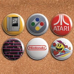Könnte beinhalten: Sechs runde Pins mit verschiedenen Retro-Videospiel-Designs. Die Pins zeigen eine gelbe und graue Nintendo-Kartusche, einen grauen Controller mit farbigen Knöpfen, ein rotes Atari-Logo, ein schwarzes und pinkfarbenes Donkey Kong-Level, ein weißes Nintendo-Logo und einen gelben Pac-Man-Charakter.