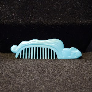 Può includere: Un pettine di plastica blu chiaro a forma di topo. Il pettine ha una fila di denti e una testa arrotondata.