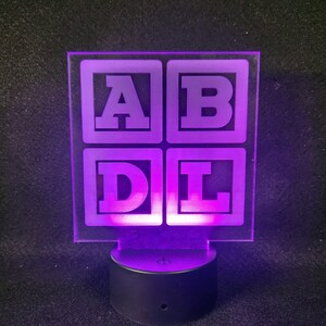 May include: A purple LED night light with a clear acrylic panel featuring four white squares with the letters A, B, D, and L inside. The night light is on a black base with a power switch.