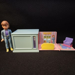 Op de afbeelding: Een blauwe en gele speelset met een pop en meubels. De speelset vouwt uit om een woonkamer te onthullen met een bank, een fauteuil en een pop die in de deuropening staat.