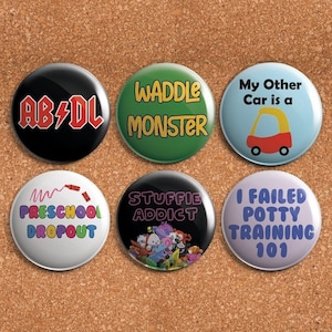 May include: Six round button pins with humorous sayings. The pins are white, black, green, blue, and purple with colorful text. The sayings include "AB/DL", "Waddle Monster", "My Other Car is a", "Preschool Dropout", "Stuffie Addict", and "I Failed Potty Training 101".